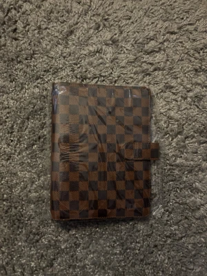 Rutigt kalenderfodral A5 - Stilrent kalenderfodral i A5-storlek med klassiskt rutmönster i varma bruna nyanser. Perfekt för dig som vill ha ordning på kalender, anteckningar eller planner – samtidigt som det ser lyxigt och tidlöst ut.  ✔ Passar A5-kalendrar / planners ✔ Smidig stängning med flik ✔ Oanvänt, fortfarande i plast ✔ Perfekt till jobb, studier eller vardagsplanering  Skick: Nytt Storlek: A5 Färg: Brun / mörkbrun rutig