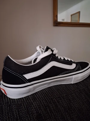Svarta Vans Old Skool sneakers - Klassiska Vans Old Skool sneakers i svart och vitt med vit sula och ikonisk sidostripe. Skorna har snörning och en platt, gummisula med det typiska rutmönstret undertill. Ovandelen är i canvas och mocka för en tidlös streetstil.