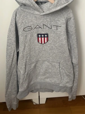 Grå hoodie från GANT med logga - Snygg gråmelerad hoodie från GANT med stor broderad logga och sköld med stjärnor och ränder på bröstet. Klassisk känguruficka framtill och huva. Perfekt för en avslappnad och sportig stil. Mjuk bomullsblandning som känns skön mot huden.