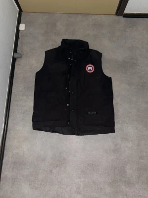 Svart dunväst från Canada Goose - Säljer en svart dunväst från Canada Goose med klassisk logopatch på bröstet och tryckknappar framtill. Västen har hög krage och två stora fickor framtill. Perfekt för lager-på-lager och riktigt snygg till höst- och vinterlooken.