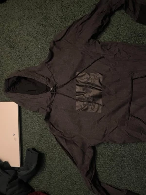 Mörkgrå hoodie från BOSS - Snygg mörkgrå hoodie från BOSS med stor svart patch och upphöjt BOSS-tryck på bröstet. Hoodien har dragsko i huvan och en klassisk känguruficka framtill. Tillverkad i mjukt bomullsmaterial som känns skönt mot huden. Perfekt för en chill och stilren look.