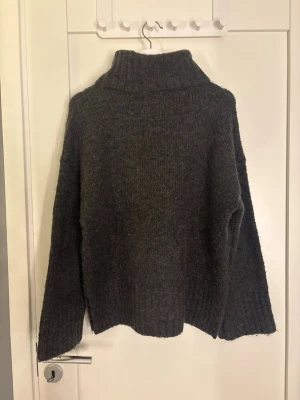 Mörkgrå stickad polotröja - Mysig mörkgrå stickad polotröja med avslappnad passform och ribbade muddar vid ärmslut och nederkant. Perfekt för lager på lager och har en hög krage som håller dig varm under kyliga dagar.