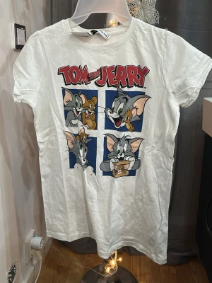 Vit Tom & Jerry t-shirt - Vit t-shirt med färgglatt Tom & Jerry-tryck framtill. Klassisk rund halsringning och korta ärmar. Perfekt för dig som gillar tecknade serier och vill ha en lekfull stil.