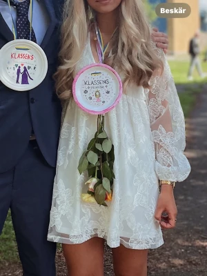 Vit blommig klännng med spets - Superfin vit kortklänning med blommigt spetsmönster och transparenta långärmar. Klänningen har v-ringning och volangdetaljer, perfekt för somriga tillfällen. Snygg och luftig passform som känns både trendig och romantisk. Superfin studentklänning som är använd endast 1 gång❣️ 