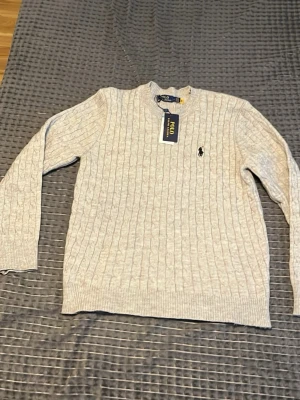 Ljusgrå kabelstickad tröja Polo Ralph Lauren - Snygg ljusgrå kabelstickad tröja från Polo Ralph Lauren med klassisk rund halsringning och den ikoniska broderade loggan på bröstet. Tröjan har ribbade muddar vid ärmslut och nederkant. Perfekt för lager på lager och stilren look.