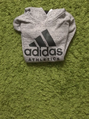 Grå Adidas hoodie med logga - Snygg gråmelerad hoodie från Adidas med stor svart logga och texten 'ATHLETICS' på bröstet. Hoodien har huva, ribbade muddar och är tillverkad i mjukt material som passar perfekt till en sportig och avslappnad stil.