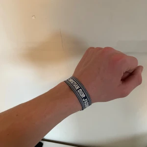 Christian Dior J'Adior tygarmband - Snyggt marinblått och vitt tygarmband från Christian Dior med texten 'J'Adior'. Armbandet har en vävd design och avslutas med fransar. Perfekt accessoar för dig som gillar exklusiva märken och vill sticka ut med en trendig detalj.