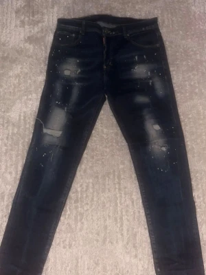 Mörkblå distressed jeans med färgstänk - Säljer ett par mörkblå jeans med slitningar och färgstänk för en riktigt edgy look. Jeansen har klassisk femficksmodell, smal passform och coola detaljer framtill. Perfekta för dig som gillar streetstyle och vill sticka ut lite extra.
