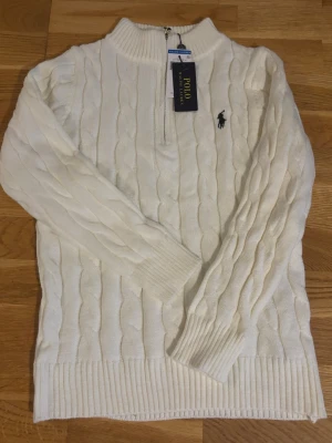 Vit kabelstickad tröja Polo Ralph Lauren - Kabelstickad vit tröja från Polo Ralph Lauren med halv dragkedja och klassisk broderad logga på bröstet. Tröjan har ribbade muddar och hög krage, perfekt för en stilren och avslappnad look. Materialet är mjukt och bekvämt.