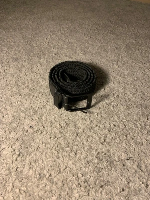 Svart flätat bälte med läderdetaljer - Snyggt svart bälte med flätad design och detaljer i läder vid spännet och änden. Spännet är i metall med matt finish och bältet har en klassisk, stilren look som passar till många outfits. (105cm långt)