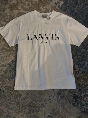 Vit t-shirt Gallery Dept x Lanvin - Säljer en clean vit t-shirt från Gallery Dept x Lanvin med tryckt logga i svart och vitt framtill. Klassisk rund hals och korta ärmar. Tillverkad i mjuk bomull som känns skön mot huden. Perfekt för dig som gillar streetwear och snygga samarbeten.