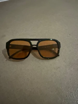 Svarta retro solglasögon med orange glas - Säljer ett par snygga retroinspirerade solglasögon med svart plastbåge och orange tonade glas. Modellen har en rak ovansida och markerad brygga, vilket ger en cool vintagekänsla. Perfekta för dig som vill sticka ut med en unik look.