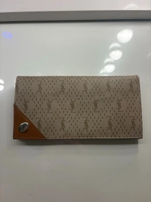 Beige YSL plånbok - Stilren beige plånbok från Yves Saint Laurent med klassiskt YSL-monogram över hela ytan. Plånboken har bruna läderdetaljer, fack för kort och sedlar samt en praktisk dragkedjeficka. Perfekt accessoar för dig som gillar lyxiga detaljer och tidlös design.