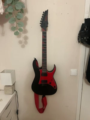 Svart och röd elgitarr med röd rem - Säljer en elgitarr i svart med röd plektrumskydd och detaljer. Gitarren har en modern, vass kropp och två humbucker-mickar. Greppbrädan har röda inlägg och tillhörande röd axelrem. Plektrum följes med. Knappt använd. Kan möta upp i Göteborg 👍🏼