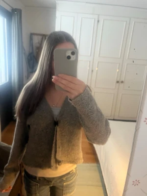 Grå croppad stickad kofta - Mysig grå croppad stickad kofta med v-ringning och knäppning framtill. Koftan har långa ärmar och en avslappnad passform, perfekt att slänga på sig över ett linne eller t-shirt.