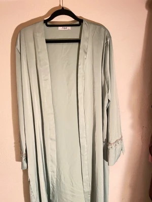Ljusgrön morgonrock med pärldetaljer - Snygg ljusgrön abaya glansigt, mjukt material. Modellen är lång med vida ärmar och har dekorativa pärlor och små kristaller längs ärmsluten för en extra lyxig känsla. Perfekt för Eid/nikkah och andra festliga tillfällen eller när du vill känna dig extra fin bara