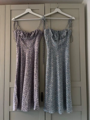 Blommiga långklänningar från Adoore - Två långklänningar från Adoore med smala axelband och söt volang vid bysten. Ena klänningen är ljusblå och den andra lila, båda med småblommigt mönster och knytband vid axlarna. Perfekta för sommarens alla tillfällen!