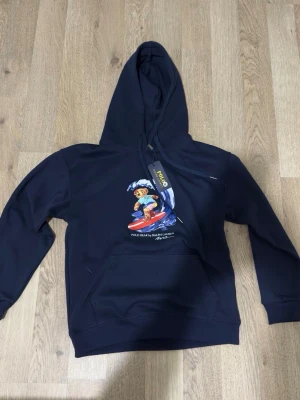 Mörkblå hoodie Polo Ralph Lauren - Snygg mörkblå hoodie från Polo Ralph Lauren med ikoniska Polo Bear som surfar på bröstet. Klassisk känguruficka framtill, ribbade muddar och justerbar huva. Tillverkad i mjukt bomullsmaterial för skön känsla. Perfekt för en avslappnad och cool stil. Storlek M men psssar även S!