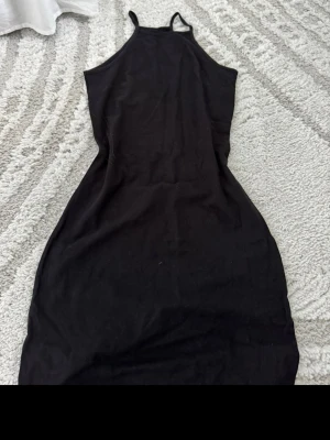 Svart ärmlös klänning med smala band - En stilren svart klänning med smala axelband och rak passform. Klänningen är ärmlös och har en enkel design som gör den lätt att matcha med olika accessoarer. Perfekt för dig som gillar minimalistisk stil.