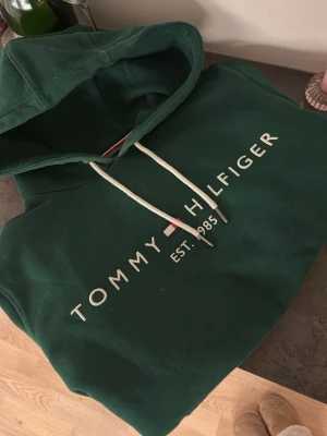 Grön hoodie från Tommy Hilfiger - Mörkgrön hoodie från Tommy Hilfiger med vit logga och text på bröstet. Klassisk huva med vita dragsnören och mjukt material, perfekt för en avslappnad stil. Snygg detalj med broderad flagga och etableringsår på framsidan.💕