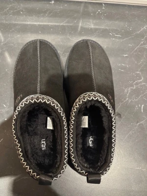 Svarta UGG tofflor i mocka - Mysiga svarta tofflor från UGG i mjuk mocka med fluffigt foder och dekorativt mönster runt öppningen. Perfekta att glida runt i hemma när du vill ha det extra bekvämt och varmt.