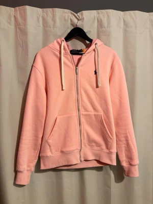 Ljusrosa zip hoodie från Polo Ralph Lauren - Ljusrosa hoodie med dragkedja från Polo Ralph Lauren. Klassisk modell med huva och vita dragsnören, två stora fickor framtill och liten broderad logga på bröstet. Tillverkad i mjukt sweatshirtmaterial, perfekt för en avslappnad stil.