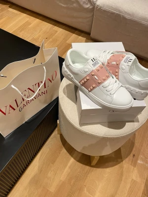Valentino Garavani sneakers med nitar - Snygga vita sneakers från Valentino Garavani med ljusrosa band och ikoniska nitar i guldton. Skorna har rund tå, platt sula och klassisk snörning. Tillverkade i skinn för en lyxig känsla och stilren look.