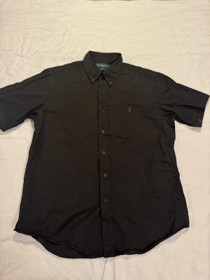 Svart kortärmad skjorta Ralph Lauren - Snygg svart kortärmad skjorta från Ralph Lauren med klassisk button-down krage och röd broderad logga på bröstet. Skjortan har custom fit och knappar framtill. Perfekt för en clean och stilren look.
