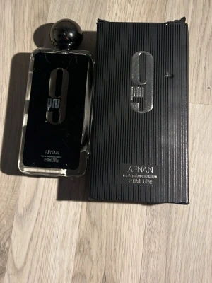 Afnan 9PM Eau de Parfum  - Afnan 9PM Eau de Parfum är en stilren herrdoft i en rektangulär glasflaska med svart etikett och silverdetaljer. Flaskan har en rund, svart kork och kommer i en matchande svart kartong med silvertext. Volymen o ml