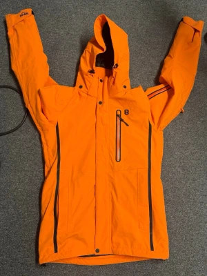 Orange vindjacka från 8848 Altitude - Säljer en orange vindjacka från 8848 Altitude med huva och svarta dragkedjor. Jackan har flera fickor, justerbar huva och detaljerade svarta linjer längs dragkedjorna. Perfekt för dig som vill sticka ut med färg och funktion.