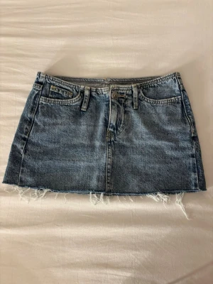 Blå kort jeanskjol - Säljer en kort jeanskjol i klassisk blå denim med rå, fransig kant nedtill. Kjolen har fem fickor, bälteshällor och knappgylf. Perfekt för en avslappnad och trendig look.