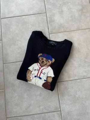 Mörkblå stickad tröja Polo Bear - Mörkblå stickad tröja från Polo Ralph Lauren med den ikoniska Polo Bear i basebolloutfit på framsidan. Rund halsringning och långa ärmar. Tröjan har detaljer i vitt, rött och blått samt RL-broderi vid nederkanten.