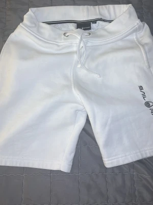 Vita Sail Racing shorts med snörning - Fräscha vita shorts från Sail Racing med snörning i midjan och tryckt logga på ena benet. Sköna att bära till vardags eller när sopranen är på topp. Tillverkade i mjukt bomullsmaterial för maximal komfort. Använt förra sommaren några gånger men inte använt sen dess 