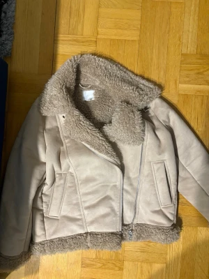 Beige pälsjacka med dragkedja - Snygg beige pälsjacka med fluffig krage och foder. Jackan har en asymmetrisk dragkedja framtill, två fickor och detaljer i samma färg. Perfekt för kalla dagar och ger en trendig look. Den är översized