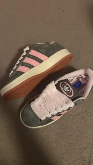 Adidas Campus sneakers grå och rosa - Säljer ett par Adidas Campus sneakers i grå mocka med klassiska rosa ränder och breda rosa skosnören. Skorna har vit sula och gummiyttersula, samt Adidas-logga på plösen. Perfekta för dig som vill sticka ut med en retro vibe.