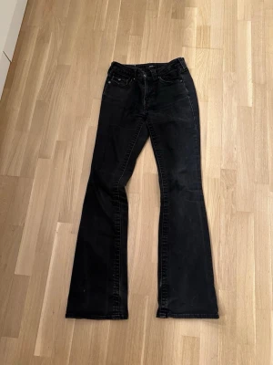 Svarta bootcut jeans från true religion  - Säljer ett par svarta bootcut jeans med snygga fickdetaljer bak och knappar på bakfickorna. Jeansen har klassisk femficksdesign och utsvängda ben.