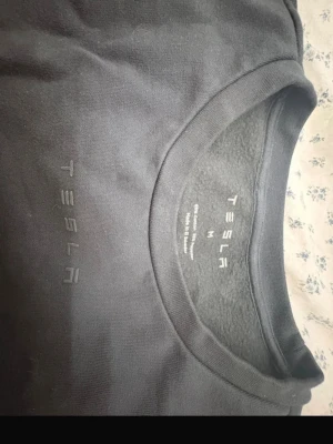 Mörkgrå sweatshirt från Tesla - Säljer en mörkgrå sweatshirt från Tesla med diskret logga på bröstet. Tröjan har rund halsringning och långa ärmar. Tillverkad i mjukt material som känns skönt mot huden. Perfekt för dig som gillar minimalistisk stil och vill representera Tesla.