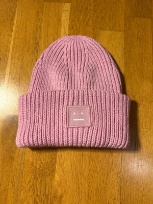 Rosa mössa från Acne Studios - Säljer en stickad rosa mössa från Acne Studios med det ikoniska ansiktslogotypen framtill. Mössan är ribbstickad och har en bred uppvikt kant för extra stilpoäng. Perfekt accessoar för att hålla värmen