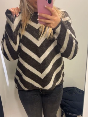 Brunvit stickad tröja med zigzag-mönster - Säljer en stickad tröja i brunt och vitt med breda zigzag-ränder. Tröjan har rund halsringning och långa ärmar. Mjuk och skön kvalitet, perfekt för kyligare dagar. Passar dig som gillar grafiska mönster och en avslappnad stil.