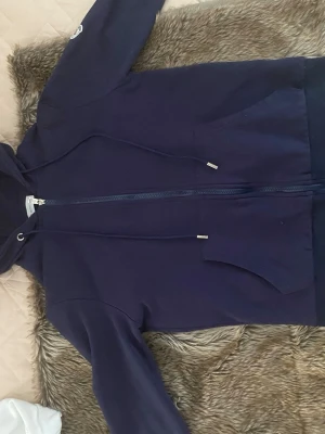 Mörkblå hoodie från Moncler - Snygg mörkblå hoodie från Moncler med dragkedja, huva och fickor framtill. Ärmsluten och huvan har detaljer i rött, vitt och blått. Moncler-logga på ena ärmen. Perfekt för en chill och sportig stil.