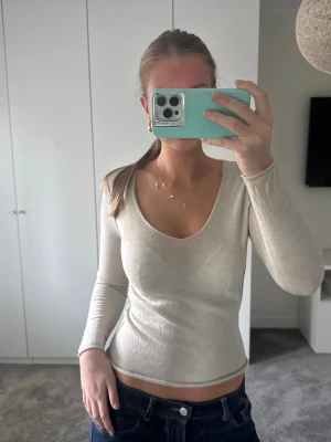 Beige långärmad topp - Trendig beige långärmad topp med djup v-ringning och snyggt åtsittande passform. På ryggen finns en stor fjärilsdetalj i silverfärgade stenar som ger en unik look. 