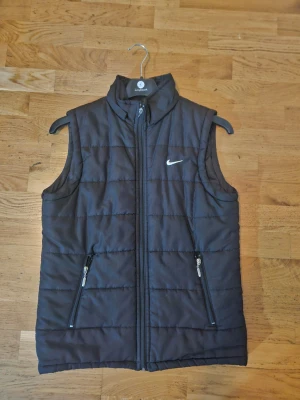 Svart dunväst från Nike - Svart dunväst från Nike med hög krage och dragkedja framtill. Västen har två snedställda fickor med dragkedja och ett diskret Nike-logga på bröstet. Perfekt för lager-på-lager och sportig stil. STORLEK S