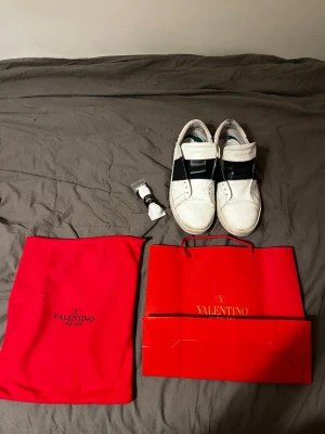 Valentino Garavani Open sneakers - Snygga Valentino Garavani Open sneakers i vitt skinn med bred svart rem över mitten. Klassisk rund tå och platt sula. Kommer med originalpåse och röd Valentino-presentpåse. Perfekta för dig som gillar exklusiva och stilrena sneakers. Den har lite skador därframme men syns inte så jätte mycket bara på nära håll. Har bild på box också men kom priv för fler frågor