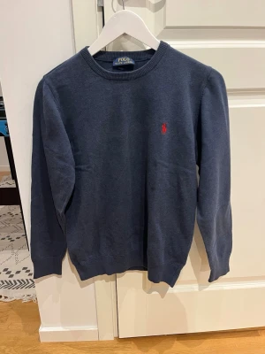 Mörkblå stickad tröja Polo Ralph Lauren - Mörkblå stickad tröja från Polo Ralph Lauren med rund halsringning och den klassiska röda logotypen broderad på bröstet. Strl S