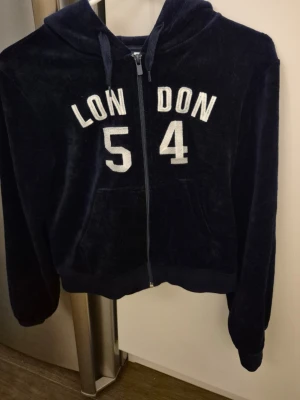 Mörkblå velourhoodie med byxor  - Mysig mörkblå hoodie i velourmaterial med dragkedja framtill och huva. På bröstet står det 'LONDON 54' i vita bokstäver och siffror. Hoodien har fickor framtill och ribbade muddar vid ärmslut och nederkant. Byxorna med fickor bak med kardborre samt snören att reglera med Som nya använda ett par gånger. OBS: Hoodie 170 cl. Byxan 158/164cl. Som XS