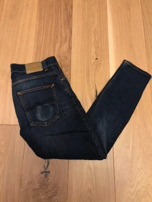 Mörkblå jeans från Nudie Jeans - Säljer ett par mörkblå jeans från Nudie Jeans med klassisk femficksdesign och kontrastsömmar. Jeansen har en rak passform och är tillverkade i slitstark bomull med Nudie-logga på bakfickan.