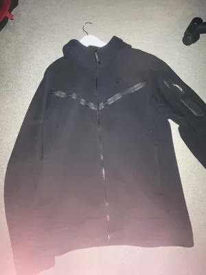 Svart hoodie från Nike med dragkedja - Svart hoodie från Nike med hel dragkedja framtill och huva. Snygg detalj med ett snedställt band över bröstet och ficka med dragkedja på ena ärmen. Perfekt för en sportig och avslappnad stil.