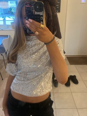 Glittrig croppad topp i silver - Säljer en glittrig croppad topp i silver med korta ärmar. Perfekt för dig som vill sticka ut och glänsa lite extra. Toppen har en rak passform och är täckt av paljetter som fångar ljuset snyggt.