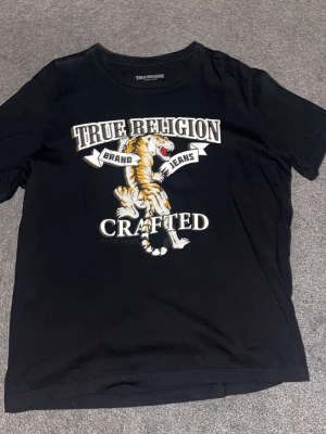 Svart True Religion t-shirt med tiger - Snygg svart t-shirt från True Religion med stort tryck av en tiger och texten 'Crafted' på framsidan. Modellen har rund hals och korta ärmar, tillverkad i mjuk bomull för skön passform. Perfekt för dig som gillar streetwear och coola prints.