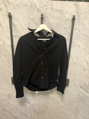 Burberry skjorta - Stilren svart skjorta från Burberry med klassiskt rutigt mönster på insidan av krage och ärmslut. Skjortan har knappar framtill och lång ärm. Det står storlek M men skulle säga att den sitter som xs den passar dig som är runt 168
